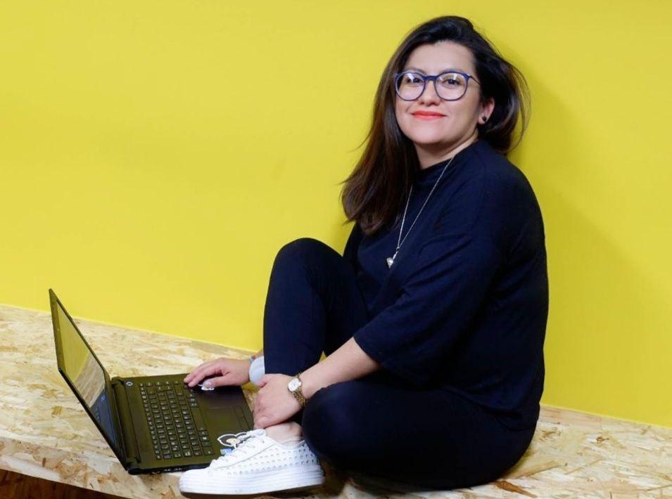 Rosa Ortega smiling and sitting in front of a laptop in a black blouse and blue glasses.